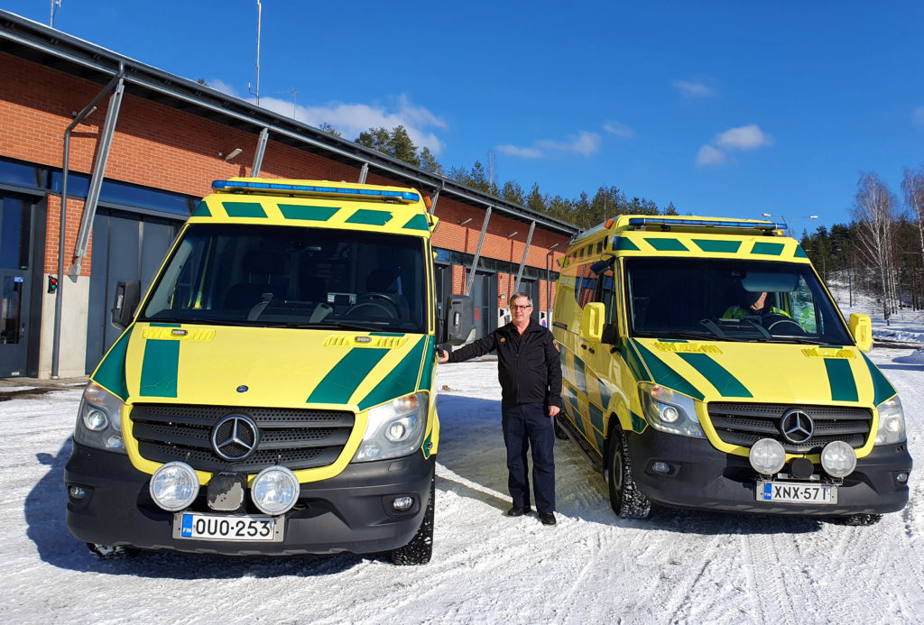 Kaksi kelta-vihreää ambulanssia pelastusaseman edessä. Niiden keskellä mies sinisessä asussa.