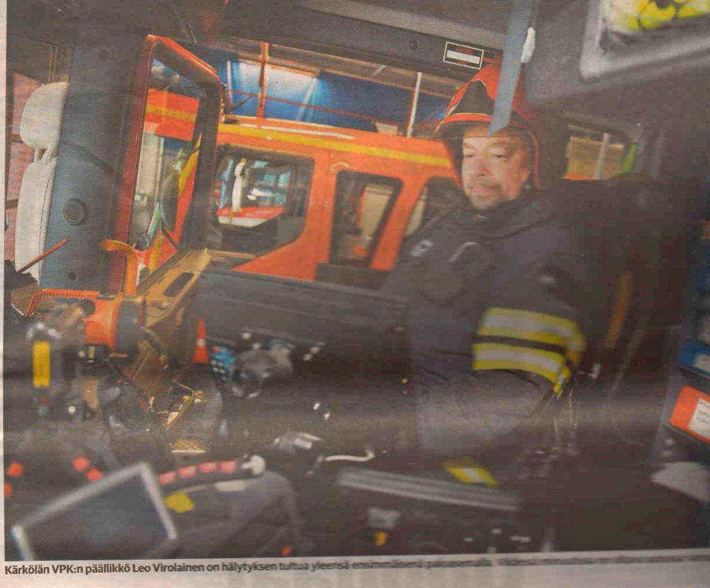 Siniseen sammutusasuun ja punaiseen kypärään pukeutunut mies keskittyneenä paloautossa istumassa. Takana toinen hälytysajoneuvo. 