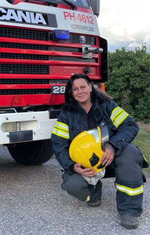 Tummahiuksinen nainen hymyilee paloauton edessä sinikeltaisssa sammutusvarusteissa keltainen kypärä sylissä. Paloautossa tunnus PH 4612.